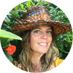 Person wearing a straw hat, smiling in a garden with green leaves and red flowers.