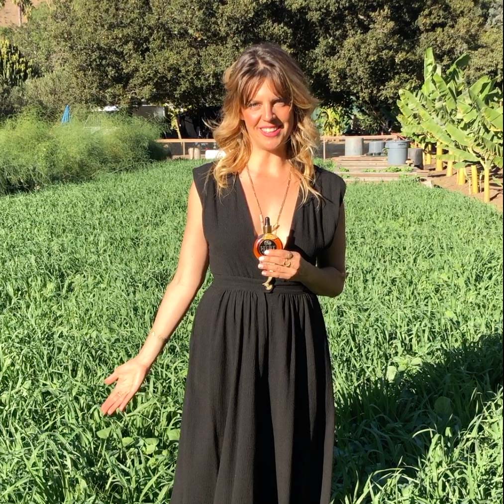 Woman in black dress holding a bottle, standing in a green field.
