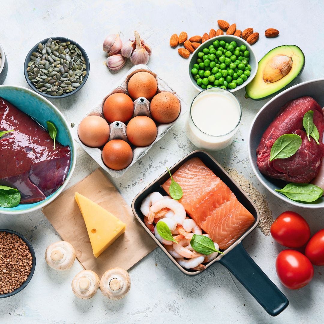 Assorted fresh foods including eggs, salmon, shrimp, avocado, and vegetables on a light background.