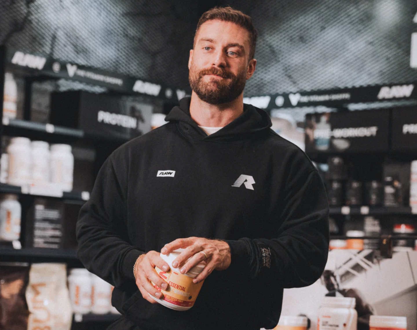 A man in a black hoodie holds a supplement container while standing in a store with shelves of products.