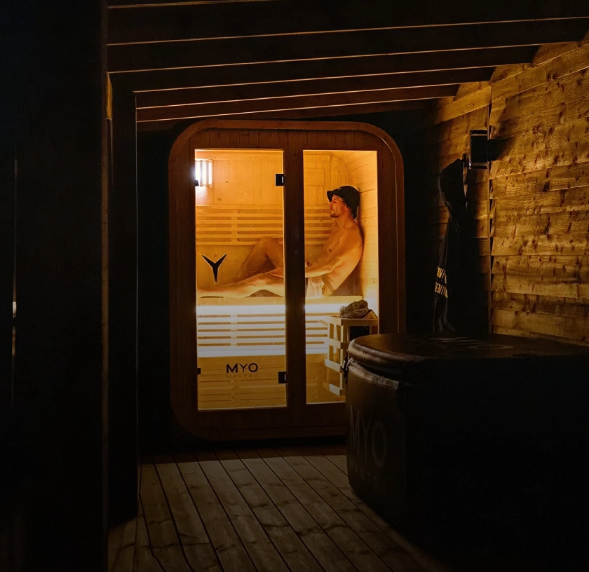 A man wearing a hat sits relaxing inside a brightly lit wooden sauna in a dark room.
