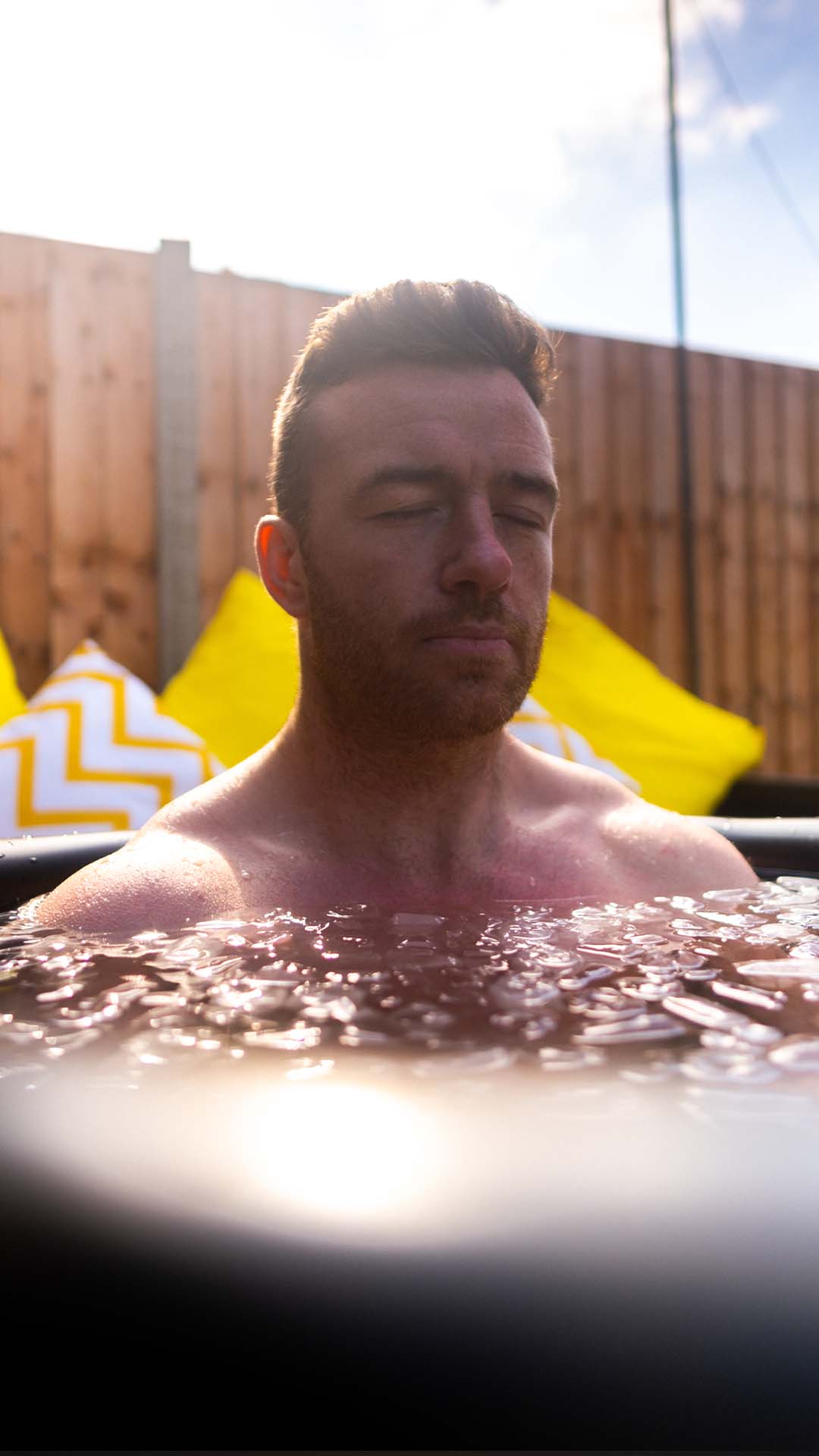 Person relaxing in a hot tub with eyes closed, wooden fence and yellow pillows in background.