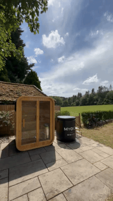 Wooden sauna and black tub on a patio with a scenic countryside view.