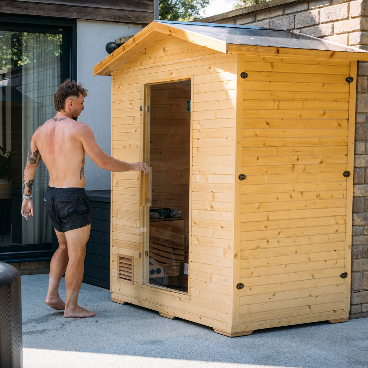 Man opening a wooden outdoor sauna with a glass door.