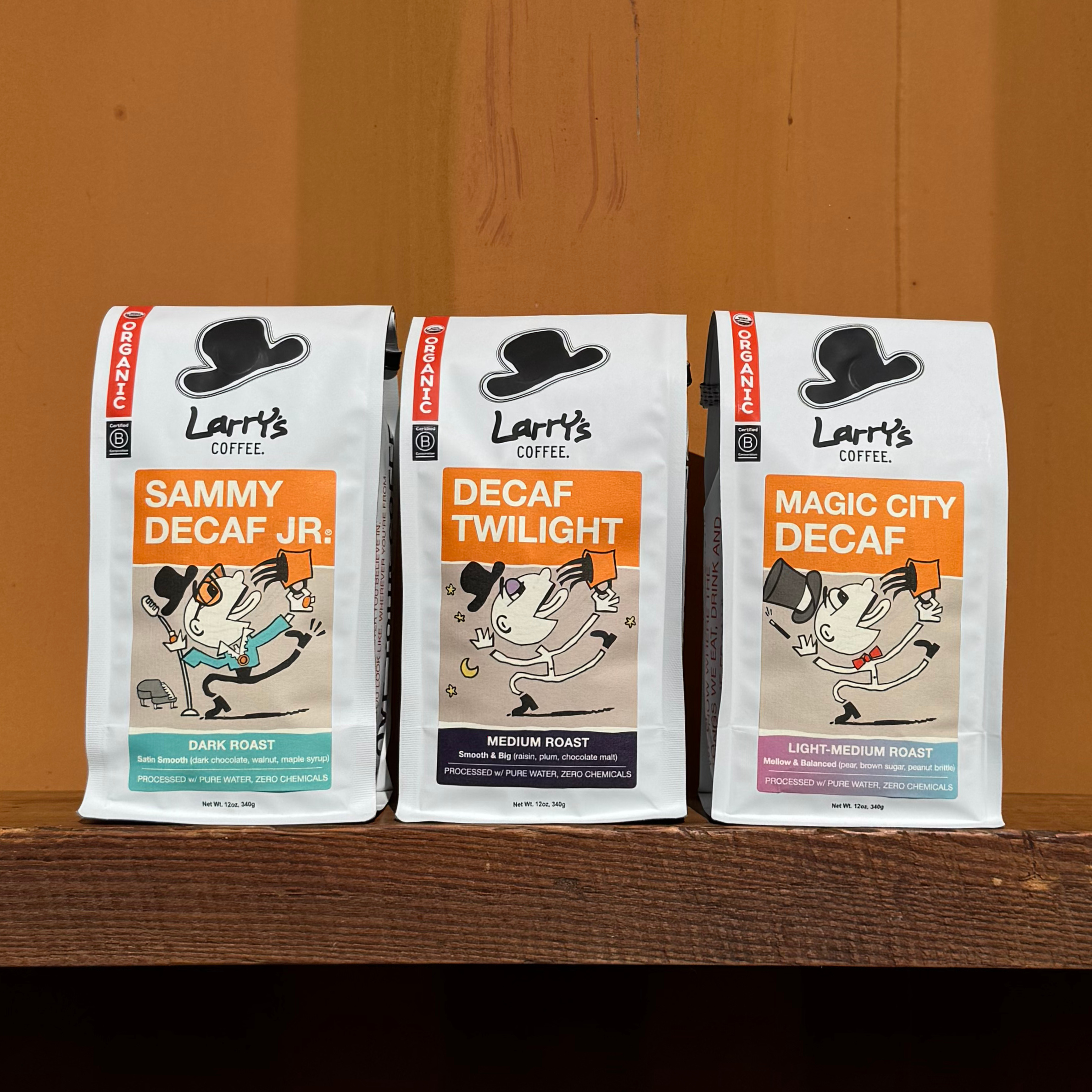 Three different bags of Larry's Decaf Coffee sitting on a wooden shelf against a tan wall.