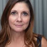 Woman with long brown hair and a neutral expression against a draped curtain background.
