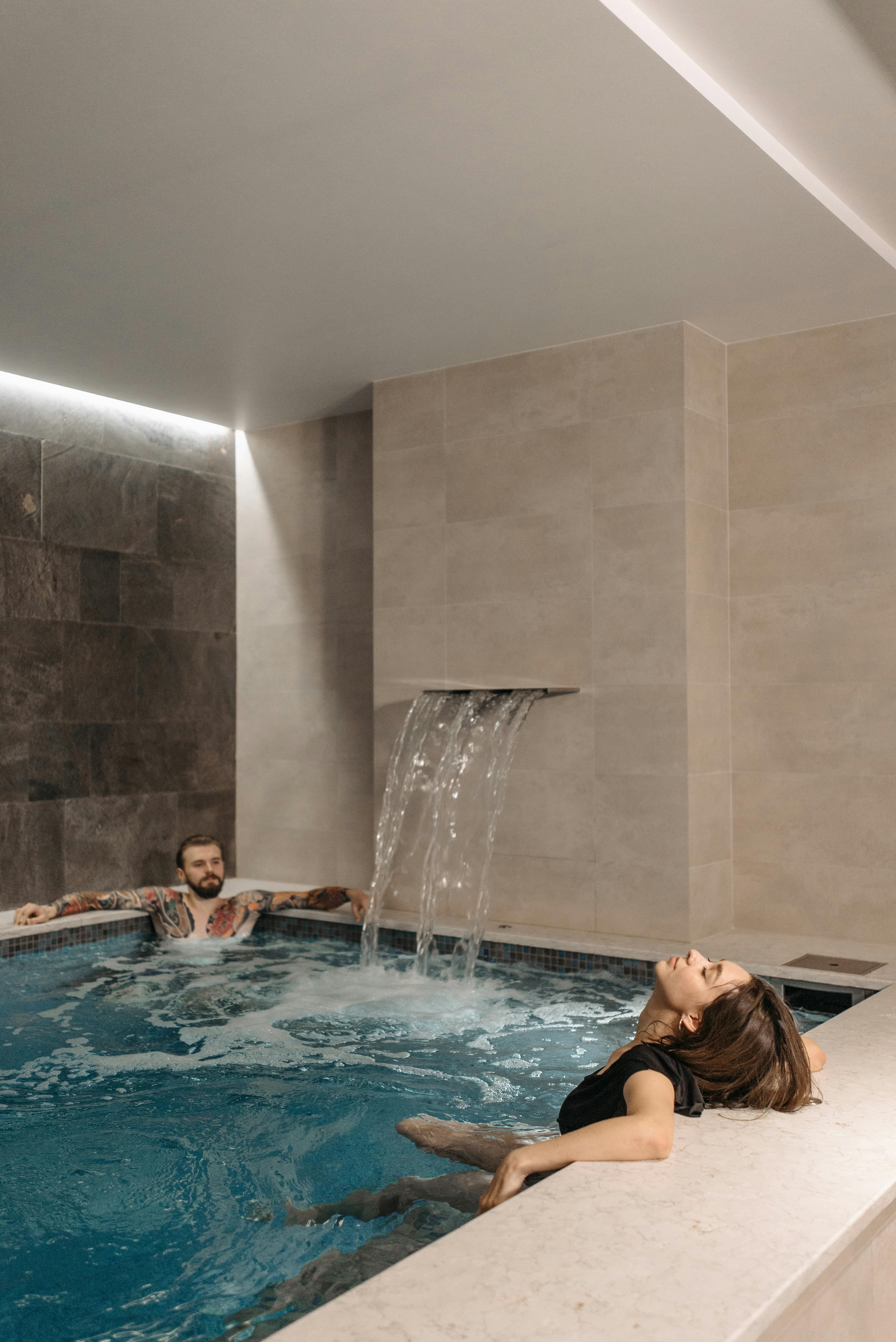 Two people relaxing in an indoor pool with a waterfall feature.