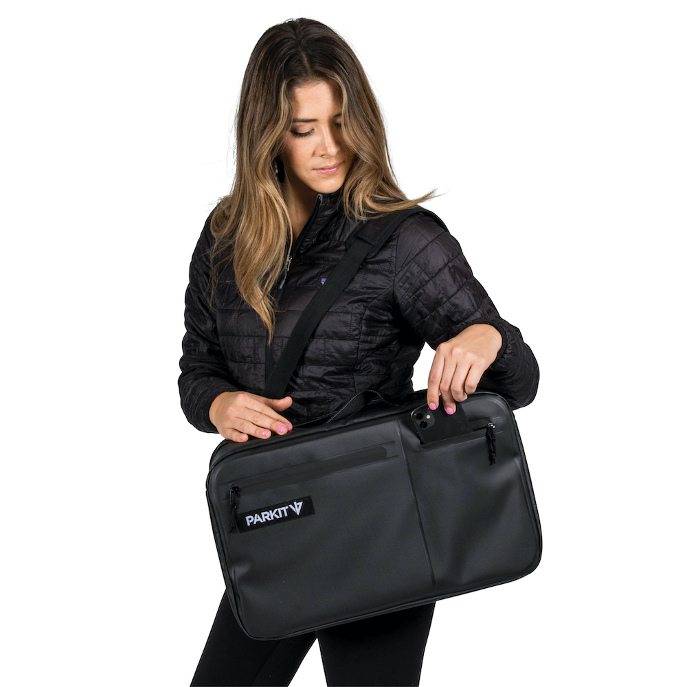 A woman in a black jacket places a phone into the outer pocket of a large black shoulder bag.