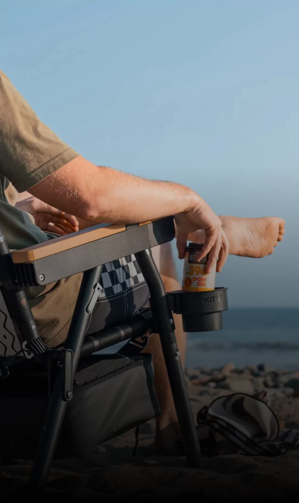 Person relaxing in a chair on the beach, holding a canned drink.