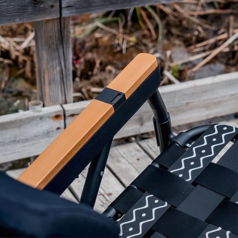 A close-up of a black chair with a tan armrest and patterned straps on a wooden deck.