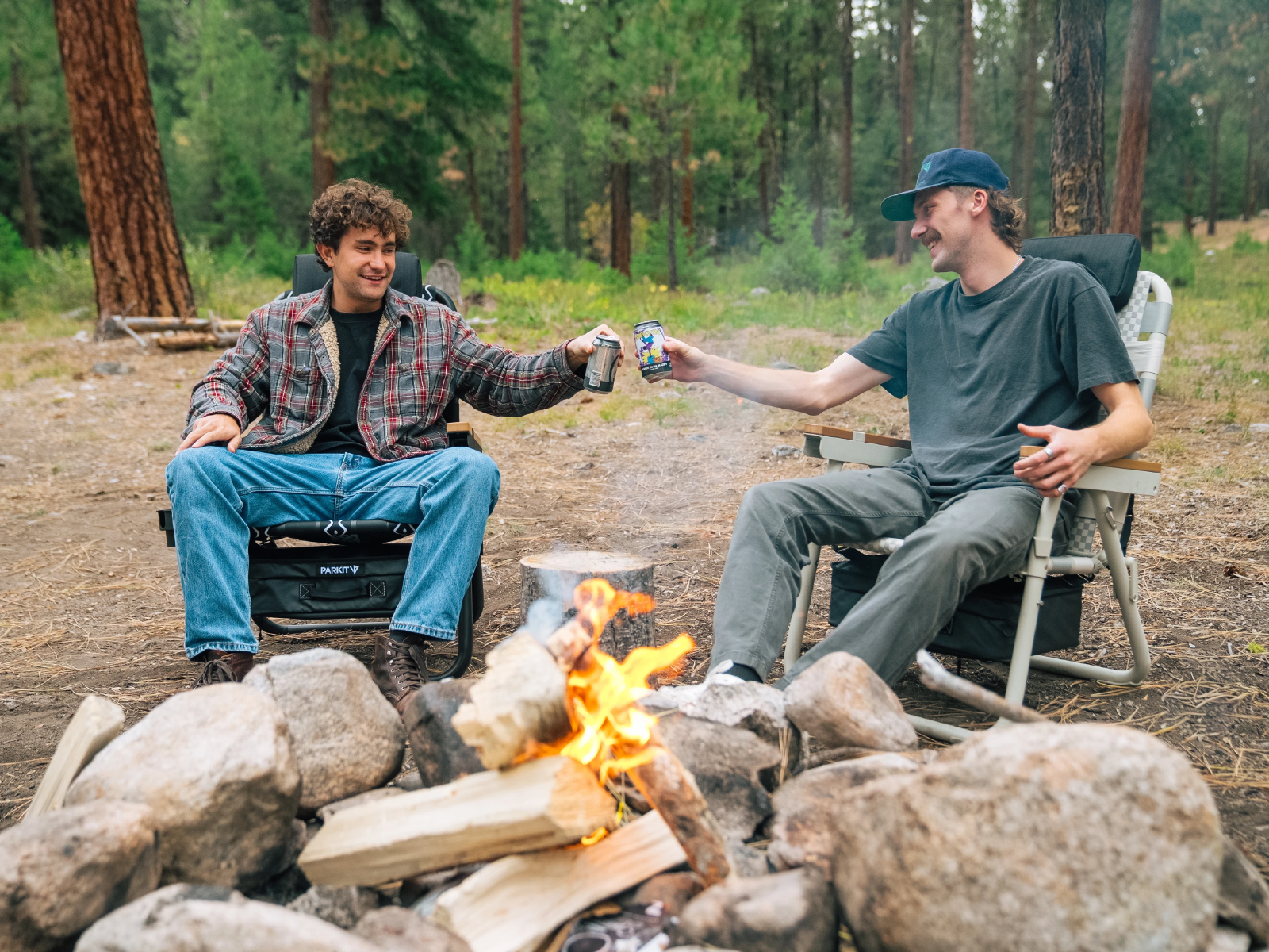 Two people sitting around a campfire, toasting with drinks in a forest.