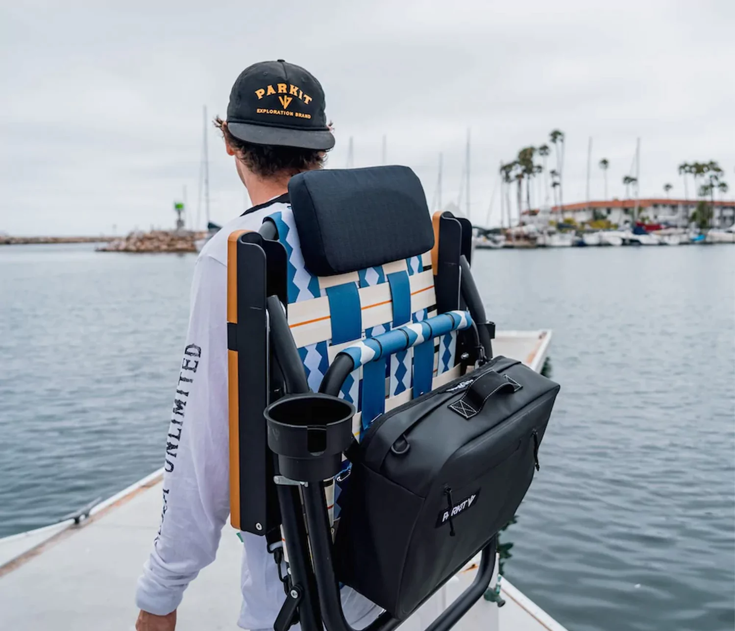 Man carrying a folded beach chair with a backpack near a marina.