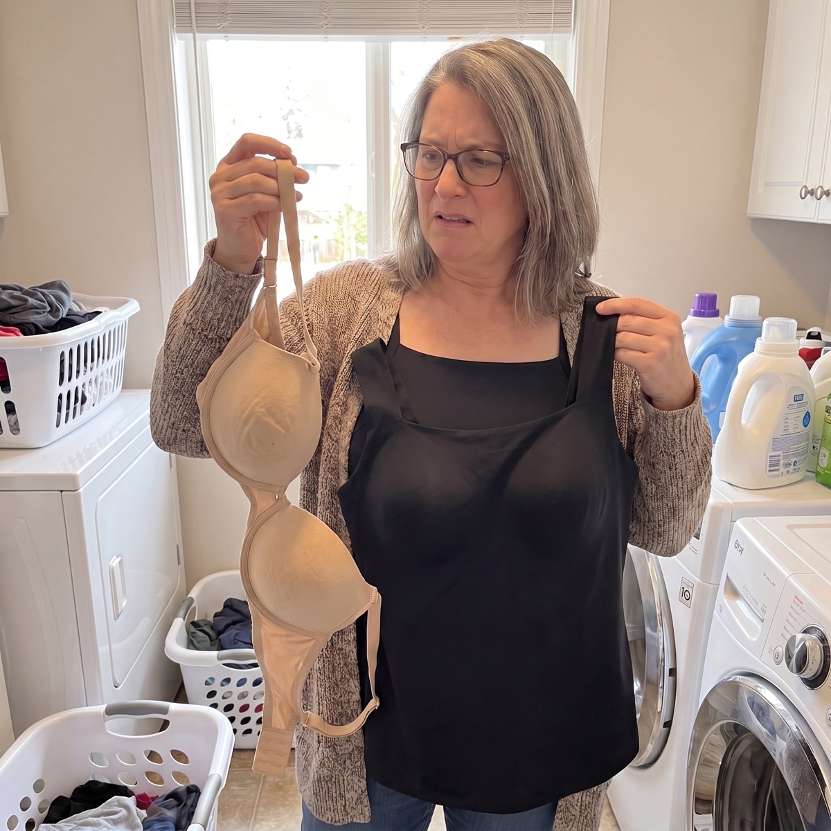 A woman in a laundry room holding up an old hook-and-eye bra in one hand and the black tank in the other