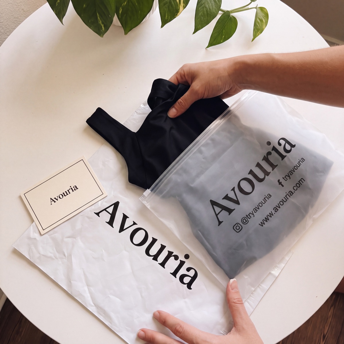 A hand pulling the black tank out of a frosted Avouria zip bag, with the Avouria mailer and thank-you card beside it