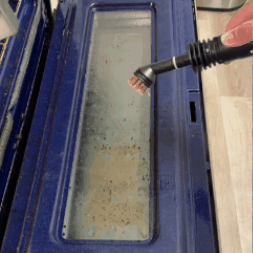 Person cleaning a dirty oven door with a brush attachment.