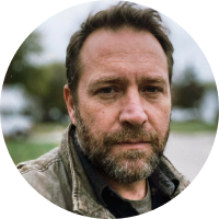 A bearded man in a jacket outdoors with blurred background.