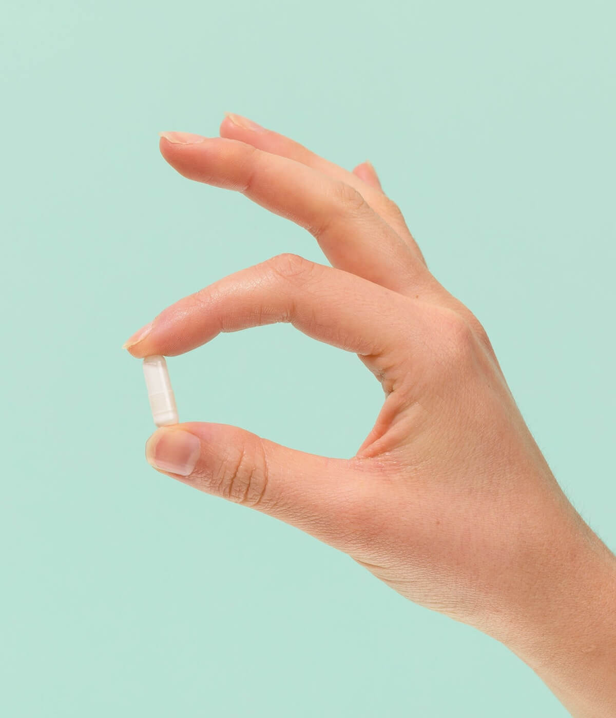 Hand holding a small capsule against a pastel green background.