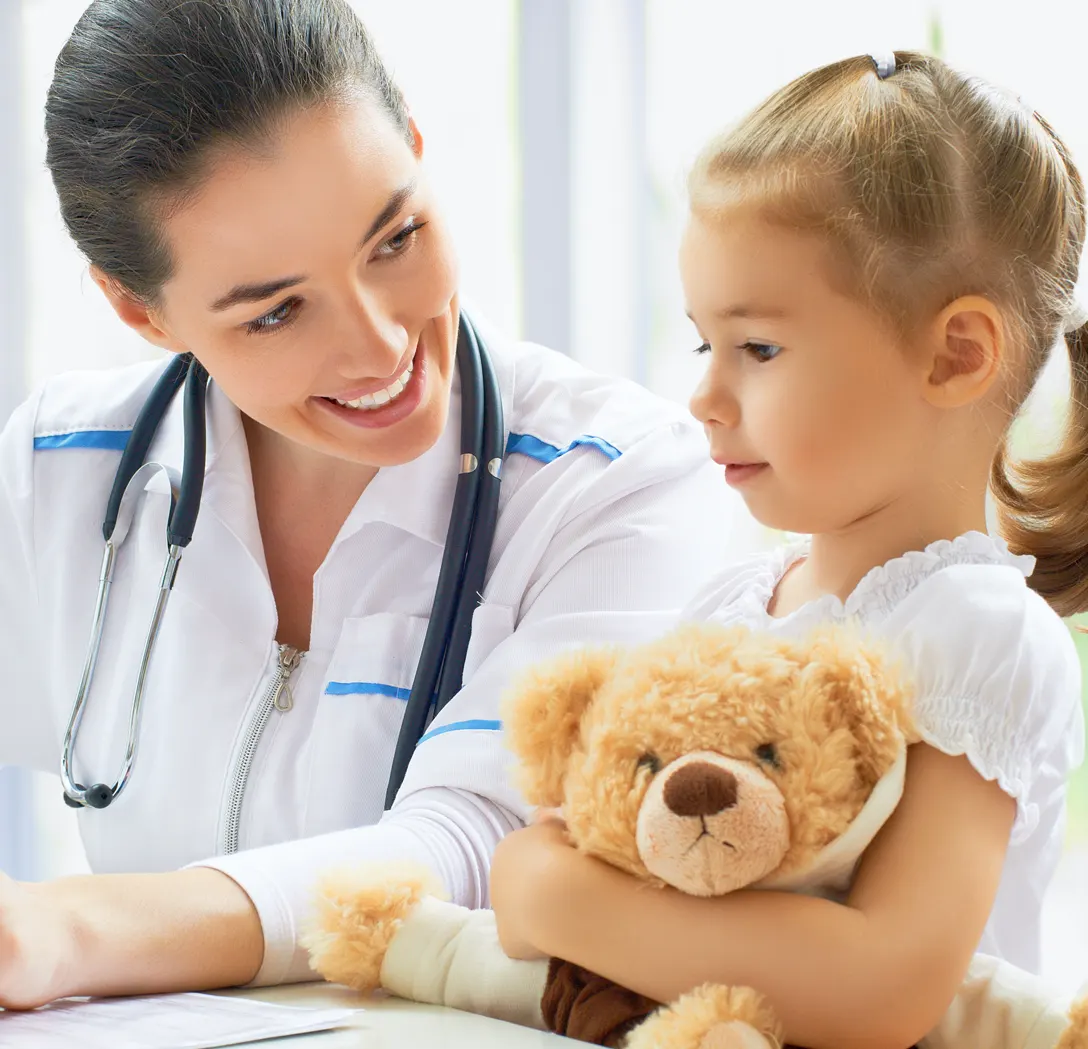 Smiling doctor with stethoscope talking to a child holding a teddy bear.