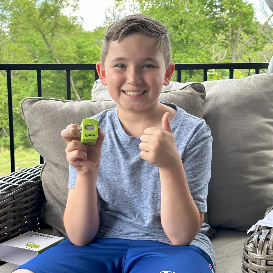 Smiling child holding a small green device, sitting on a patio with greenery in the background.