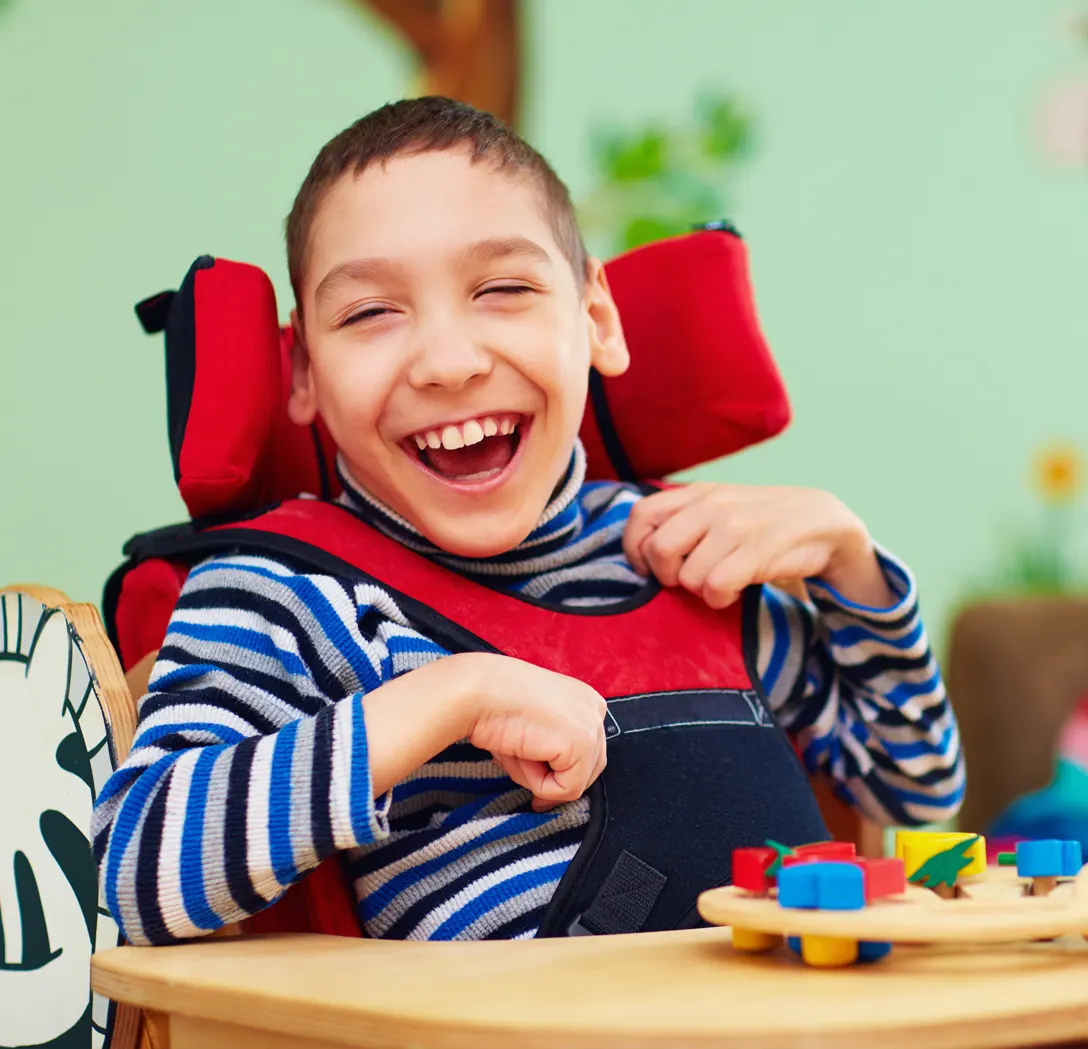 Smiling child in a striped shirt sitting in a chair with colorful toys on a table.