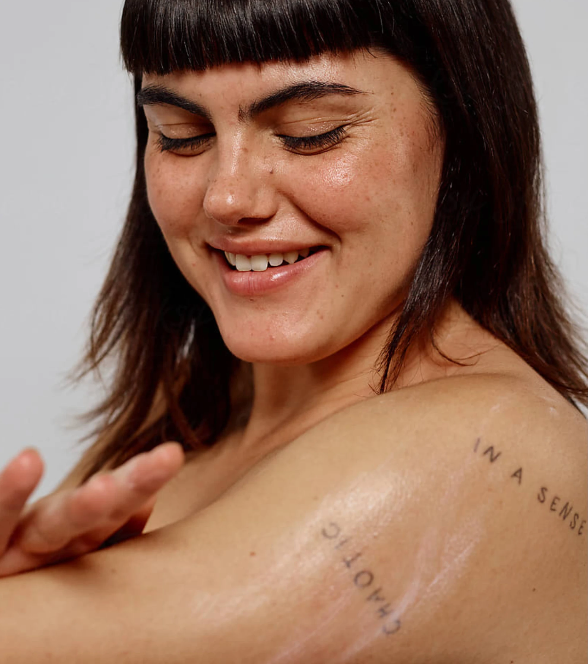 A close-up of a smiling woman with bangs and freckles applying lotion to her tattooed shoulder.