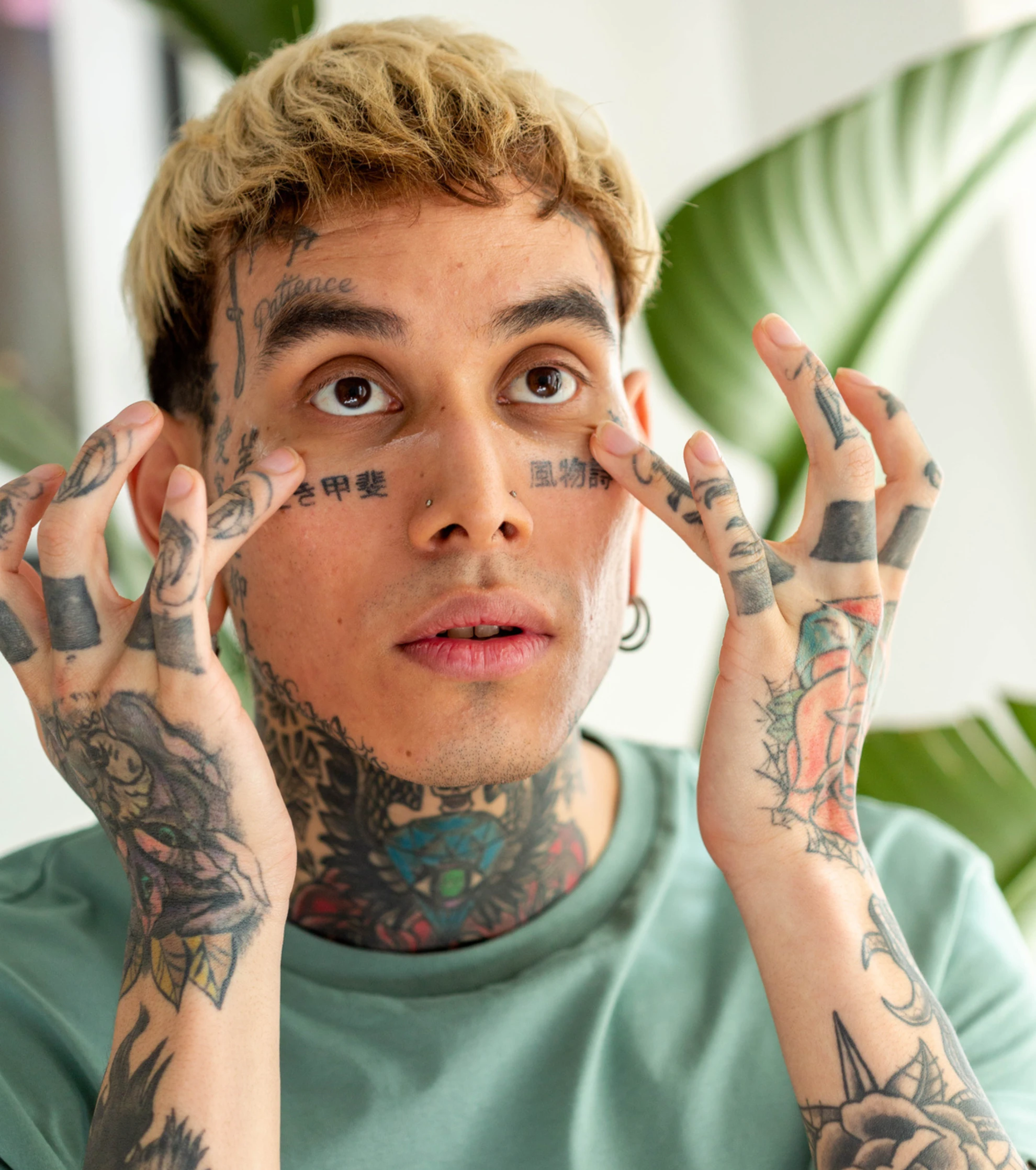 A heavily tattooed person with blonde hair looks up while touching the skin under their eyes.