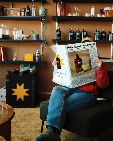 Person reading a newspaper in a cozy room with shelves of bottles.