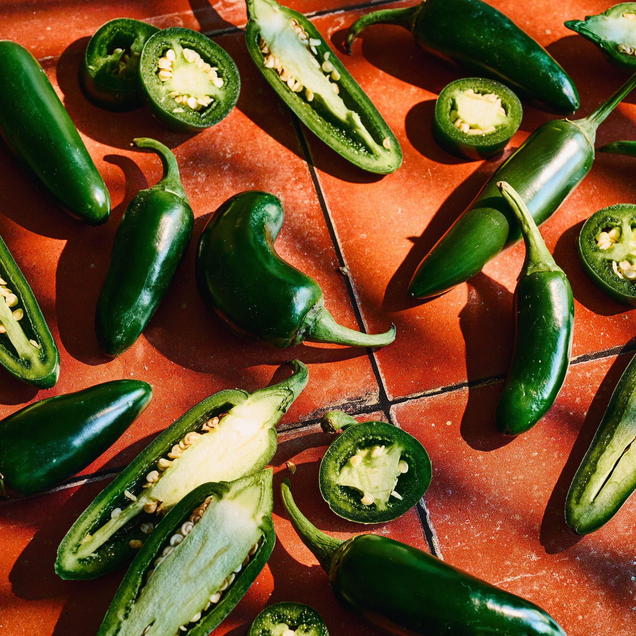 Whole and sliced green jalapeño peppers scattered on a red-orange tiled surface in harsh sunlight.