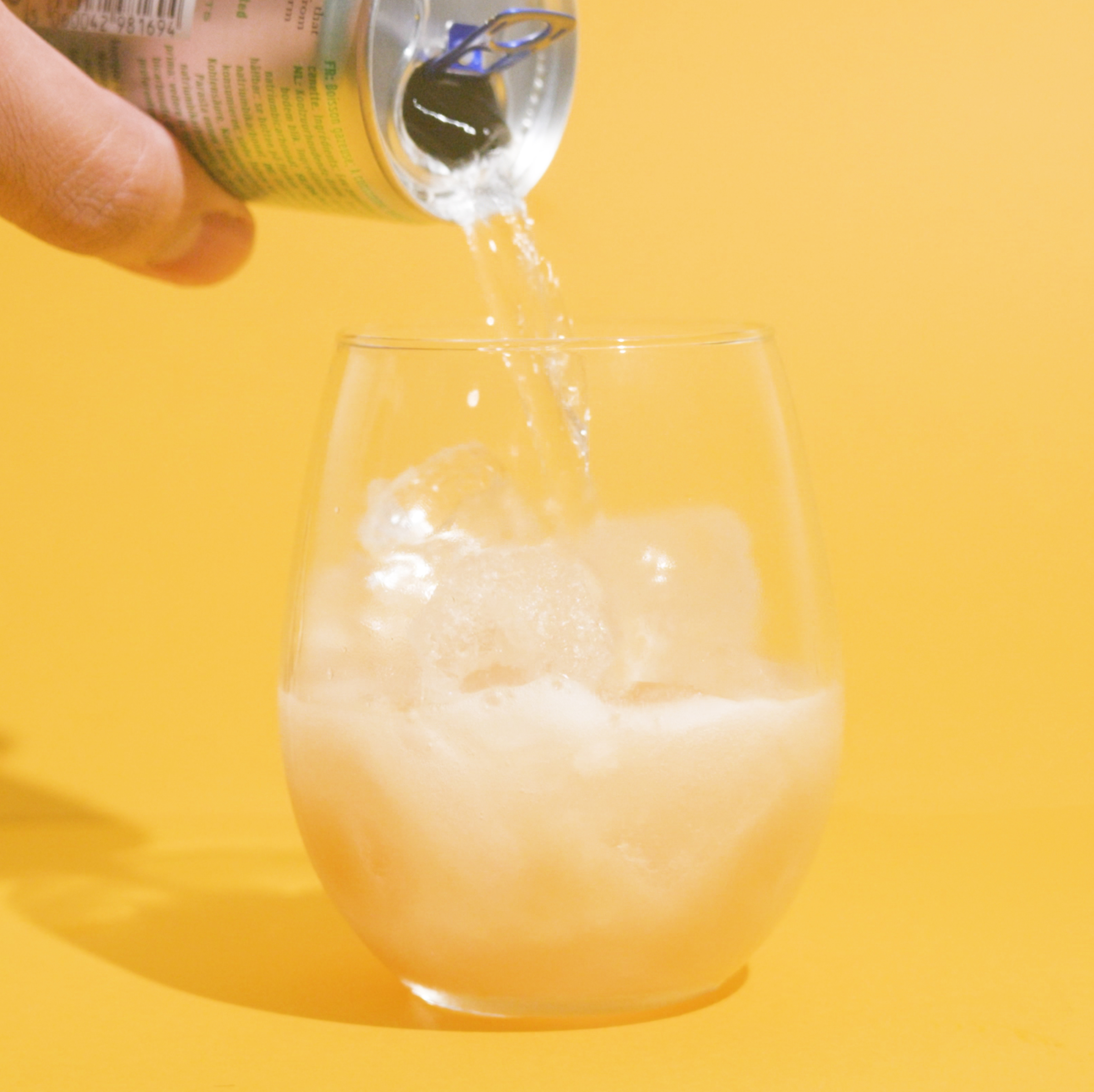 A can pouring liquid into a glass with ice against a yellow background.