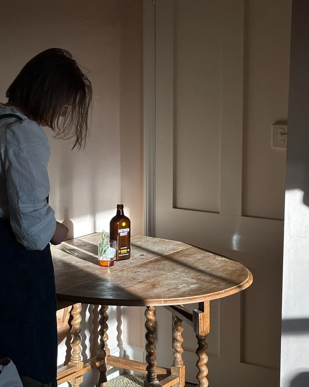 Person standing by a wooden table with a bottle and glass.