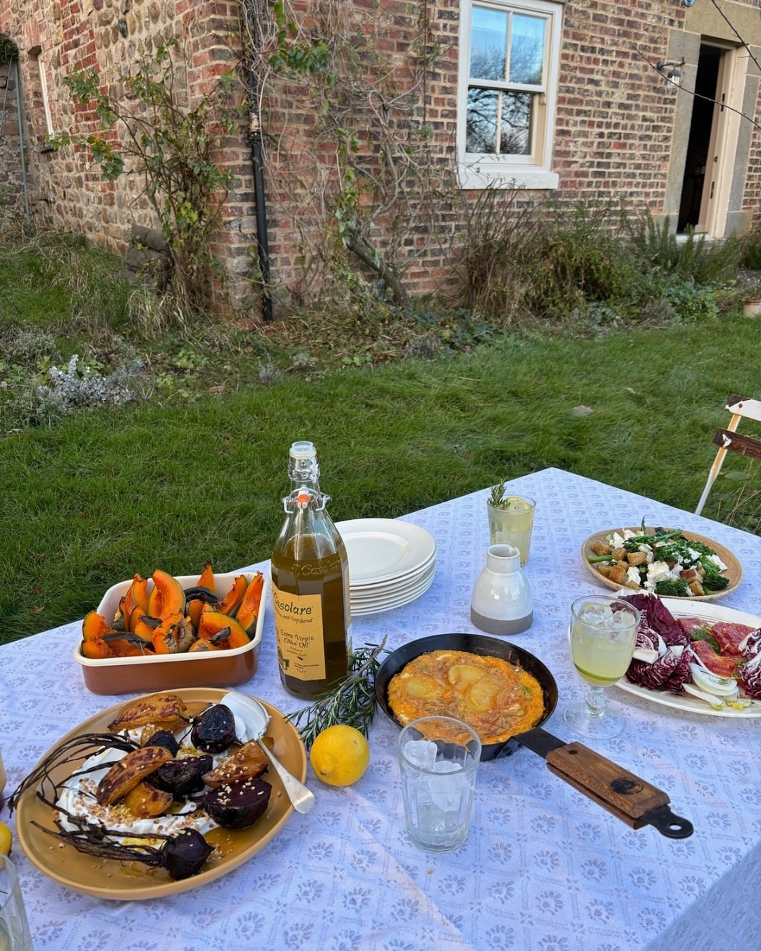 Outdoor table with various dishes, drinks, and a brick house in the background.