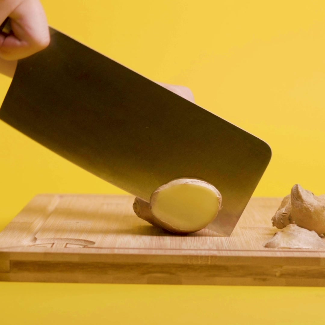 A cleaver slicing ginger on a wooden cutting board against a yellow background.