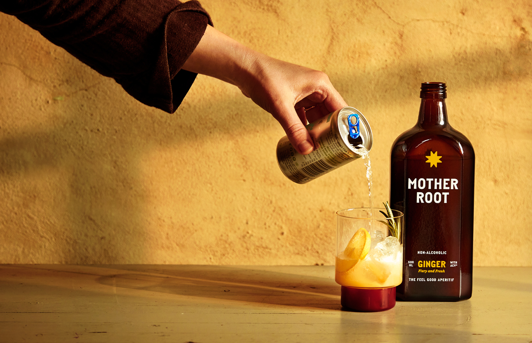A hand pours liquid from a can into a glass beside a Mother Root bottle.
