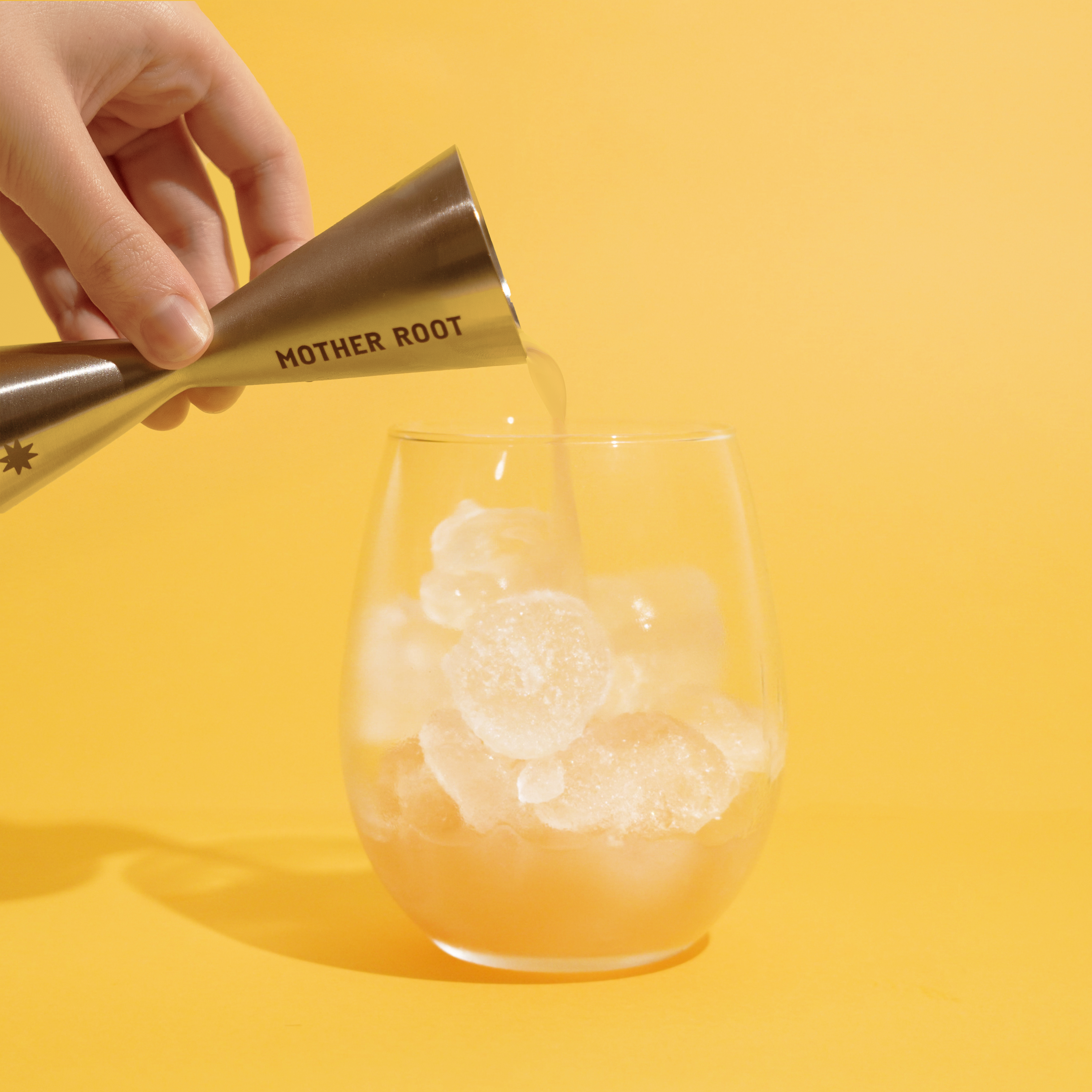 Hand pouring liquid from jigger into glass with ice, yellow background.