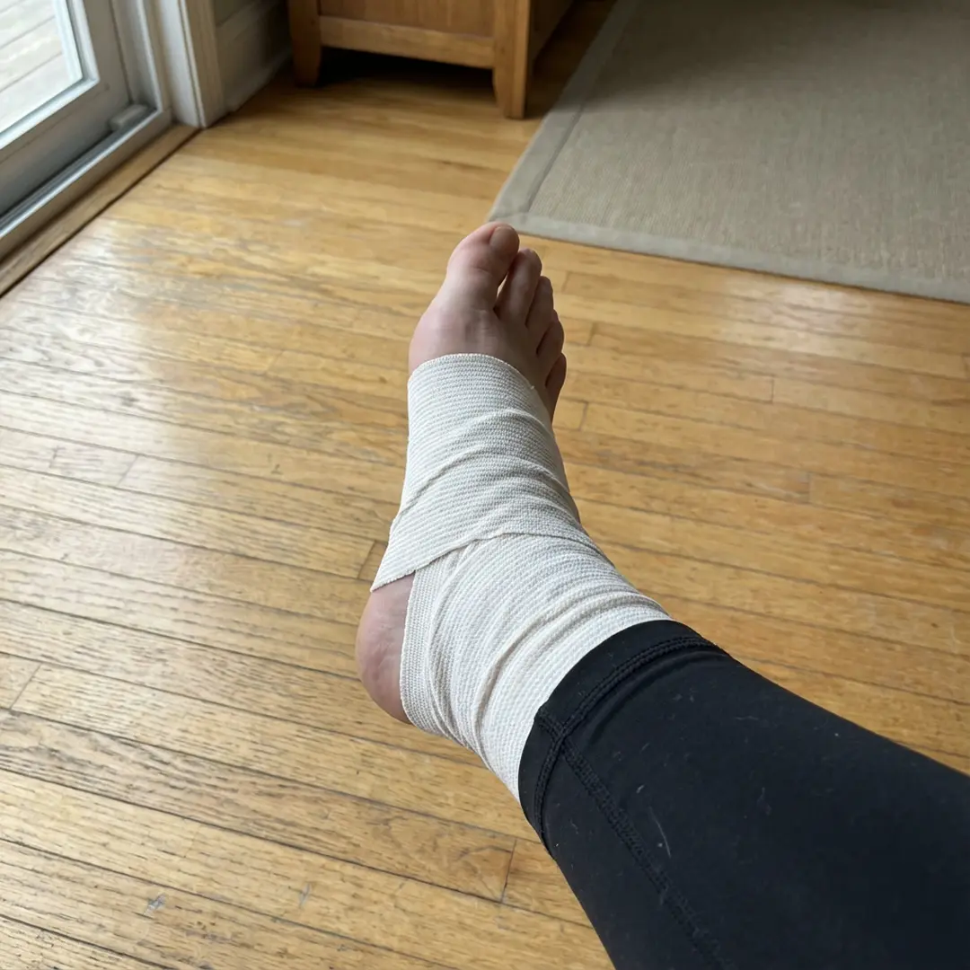 Foot with an ankle bandage resting on a wooden floor.