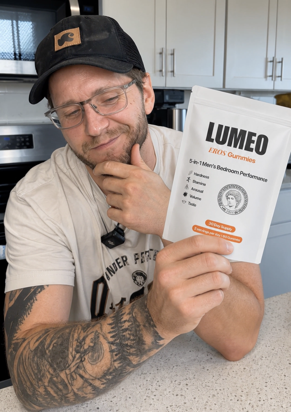 A man with glasses and tattoos holds a white pouch of Lumeo Eros Gummies in a kitchen.