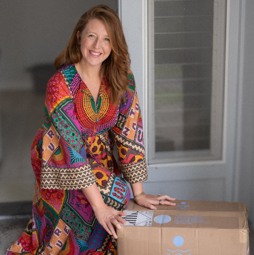Person in colorful dress smiling, kneeling beside a large cardboard box.