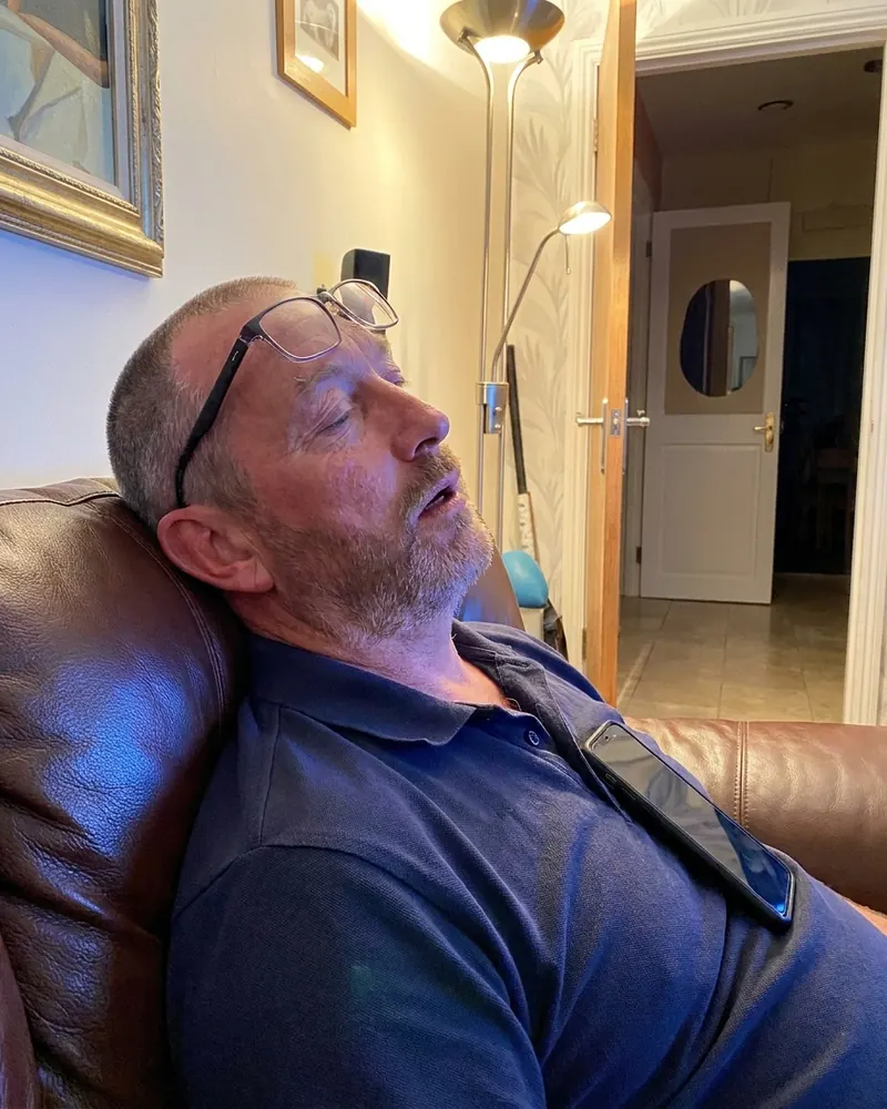 The same man asleep on a leather couch after dinner, phone on his chest