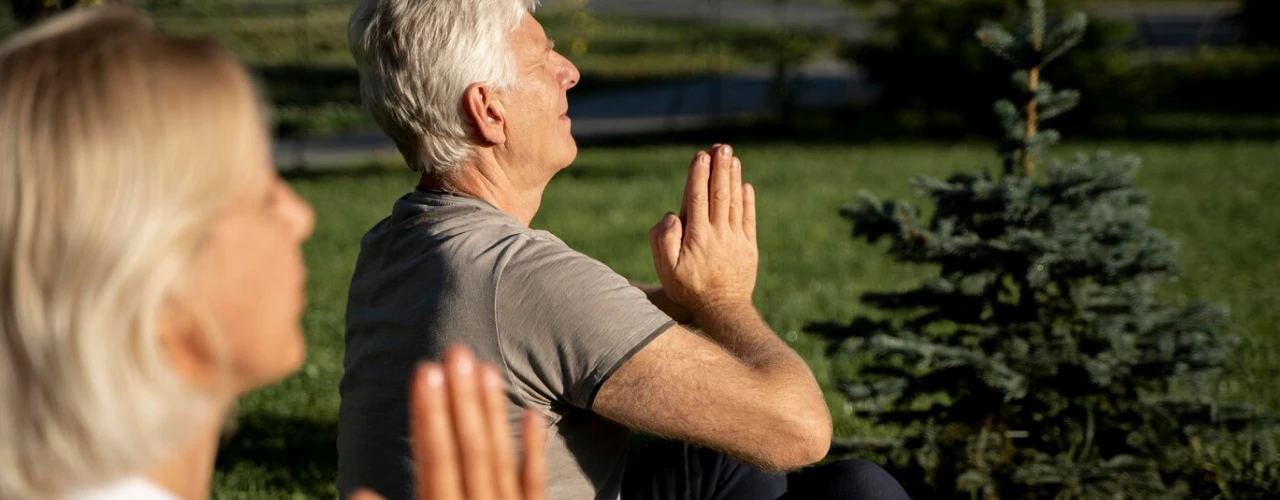 Two people meditating outdoors in a park, sitting cross-legged with palms together.