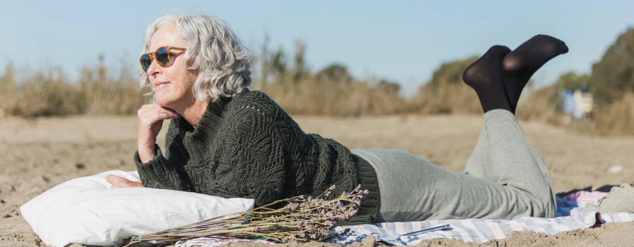 Woman with gray hair lying on a blanket at the beach, wearing sunglasses and holding lavender.