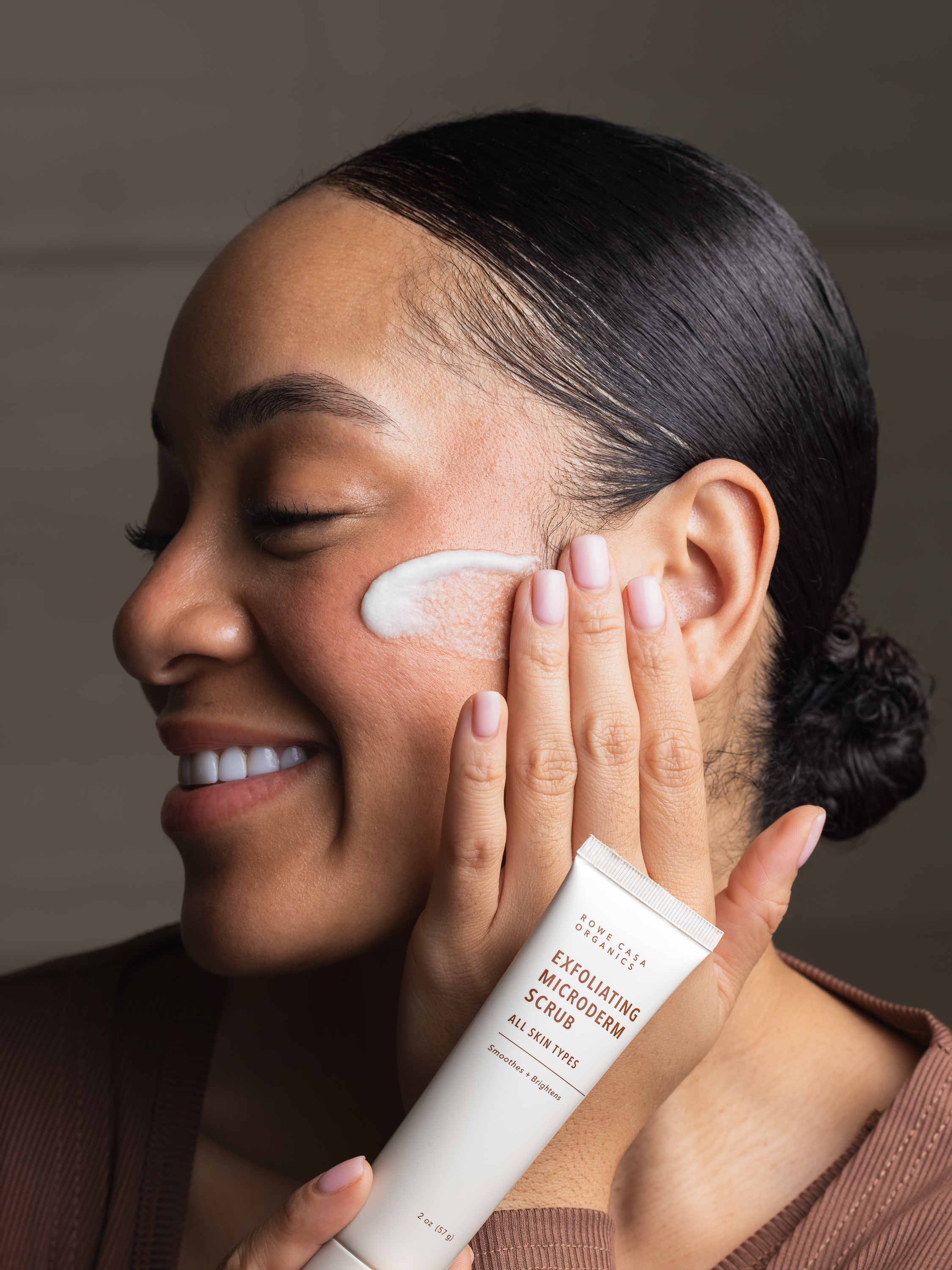 A smiling woman with her hair pulled back applies an exfoliating microderm scrub to her cheek.