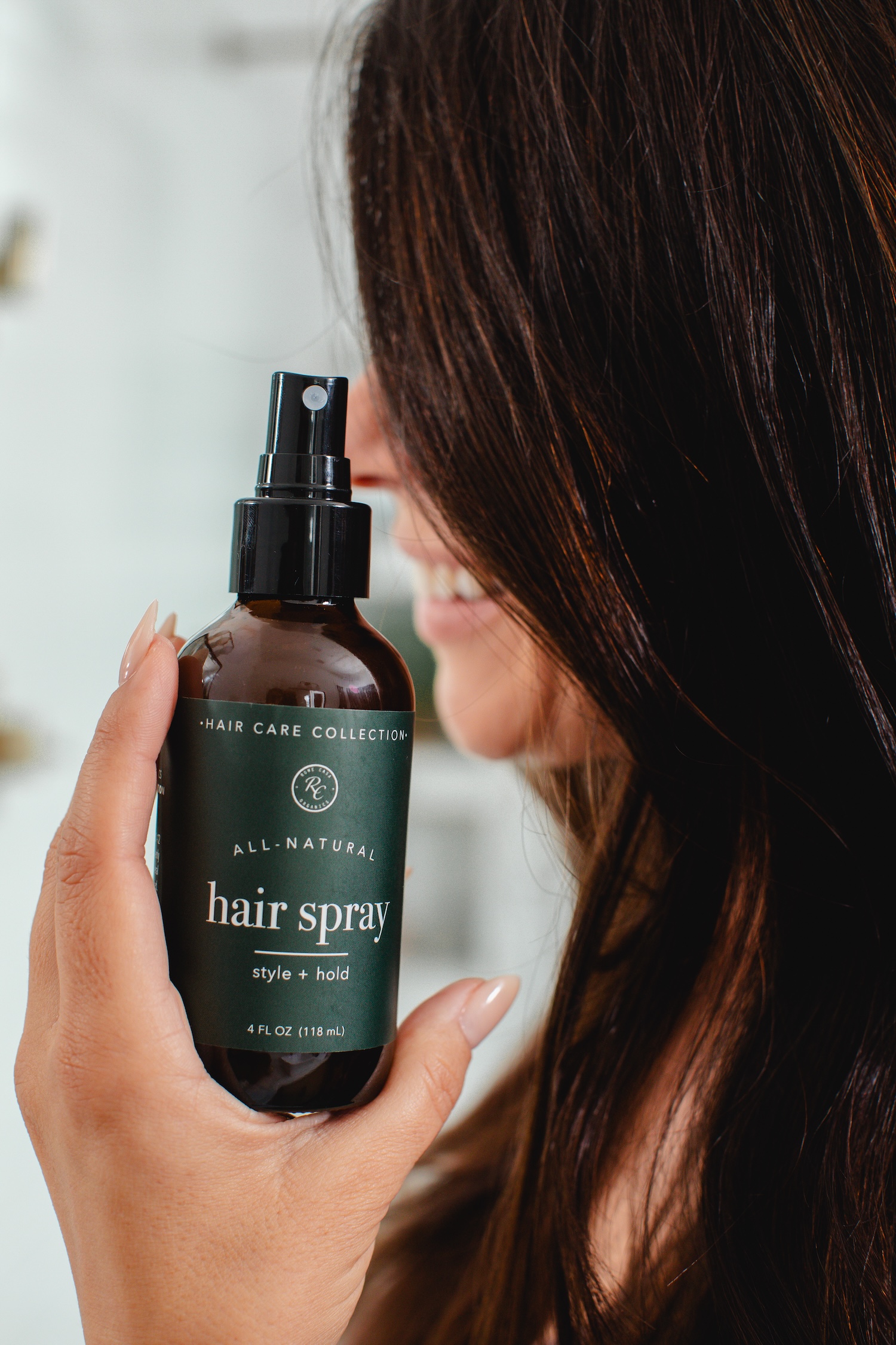 A woman with long dark hair holds a brown bottle of all-natural hair spray near her face.
