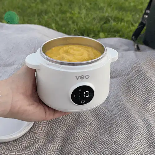 A hand holding a white, portable Veo food container filled with yellow puree on a picnic blanket.
