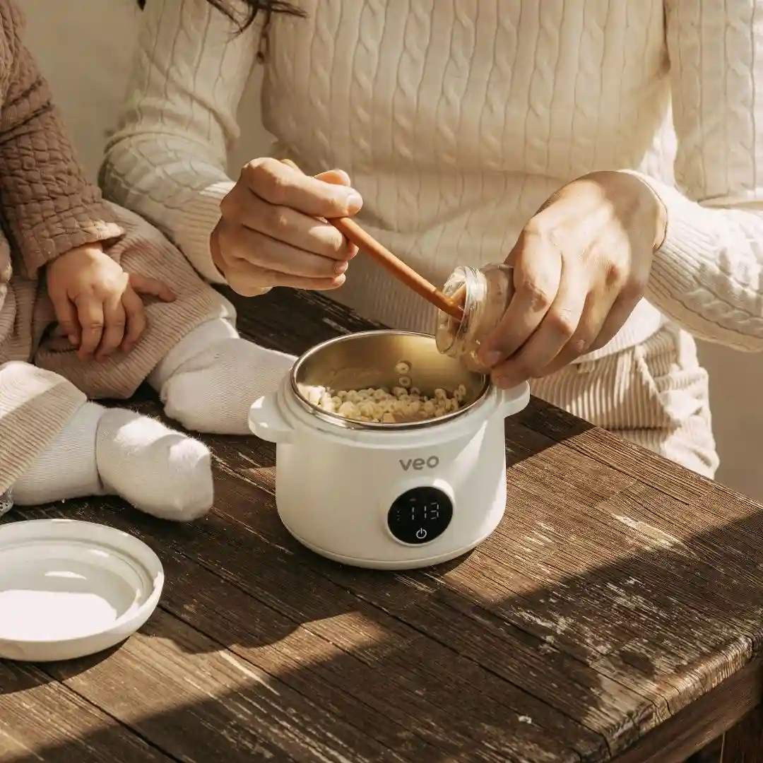 A person adds ingredients to a small, white electric pot while sitting next to a baby.
