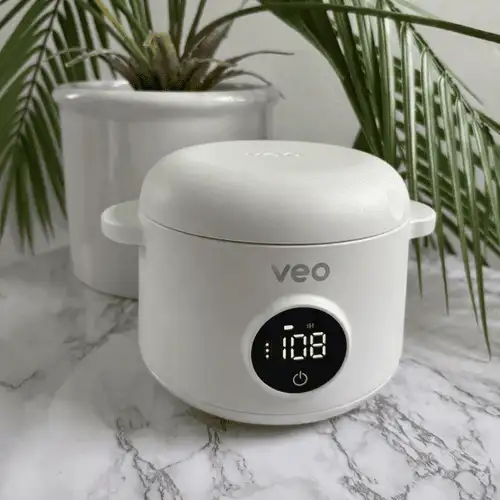 A small white Veo electric cooker with a digital display sits on a marble countertop near a plant.