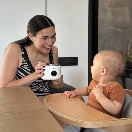 A woman smiles as she holds a small food warmer in front of a baby in a highchair.