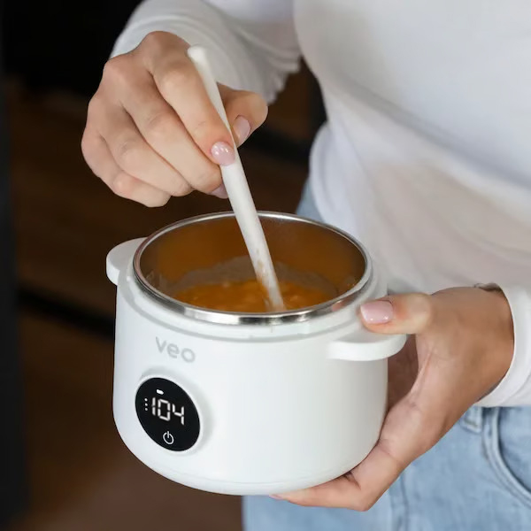 A person stirs an orange-colored substance in a small, white electric pot with a digital display.