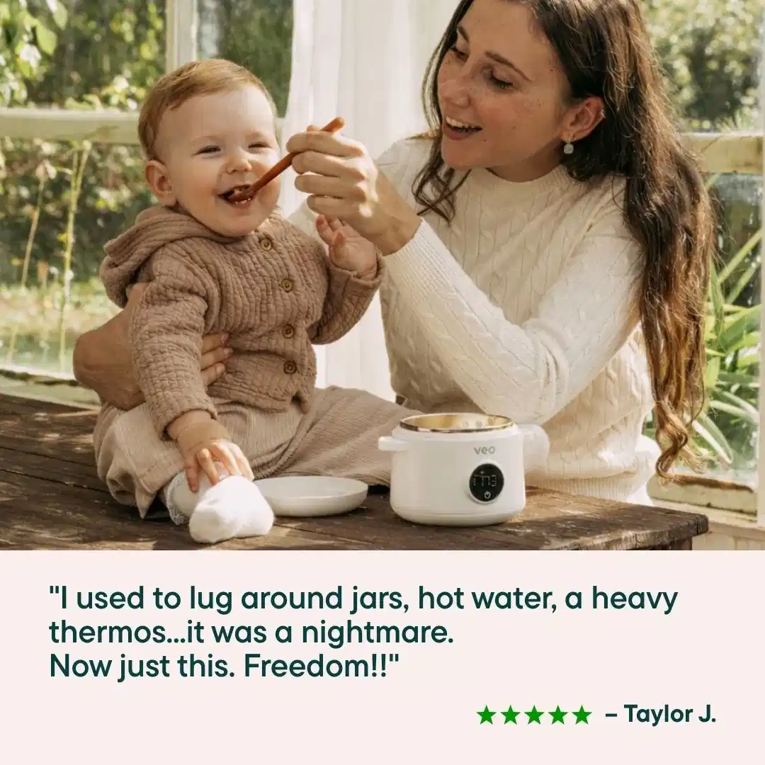 A smiling woman spoon-feeds a happy baby sitting on a wooden table next to a small white appliance.