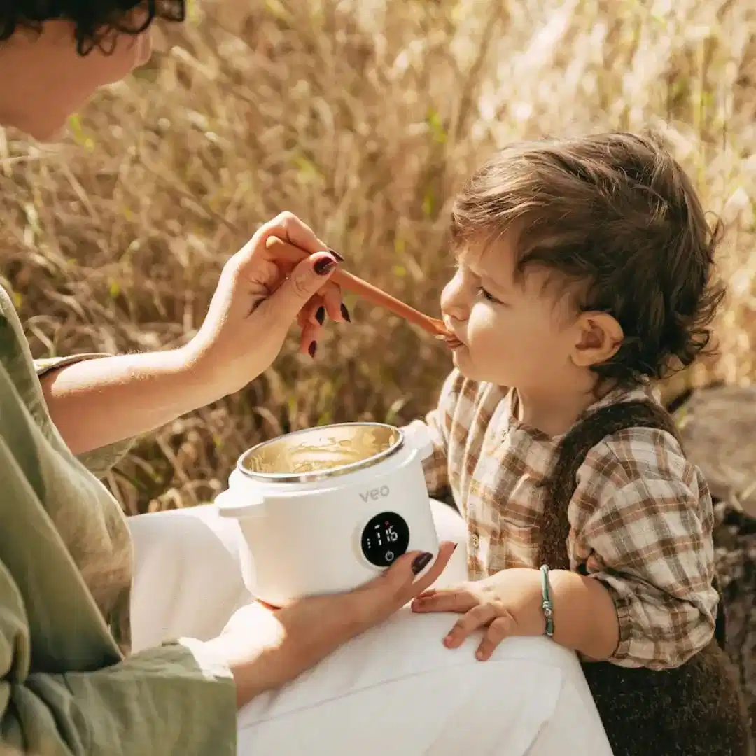 A woman feeds a toddler with a spoon from a portable food warmer while sitting outdoors.