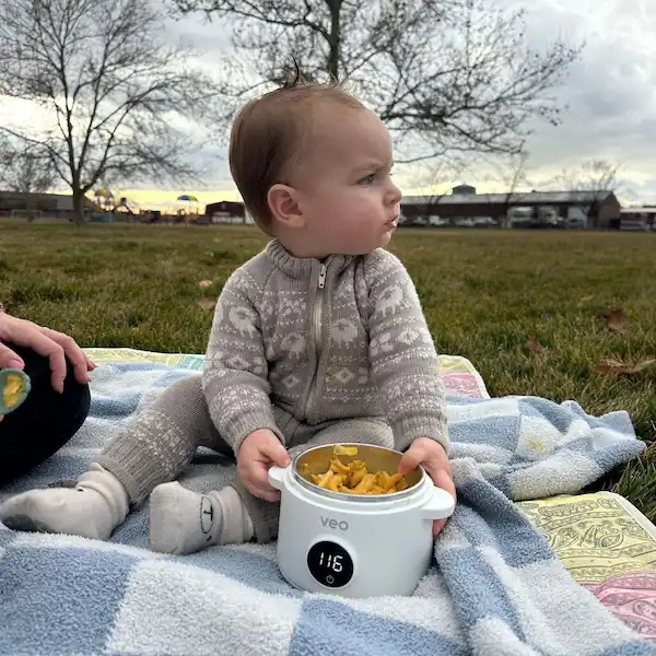 A baby in a knit romper sits on a blanket in a grassy park holding a food warmer.